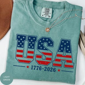 A light green t-shirt with the text "USA" and "1776-2026" printed on it, along with an American flag design.