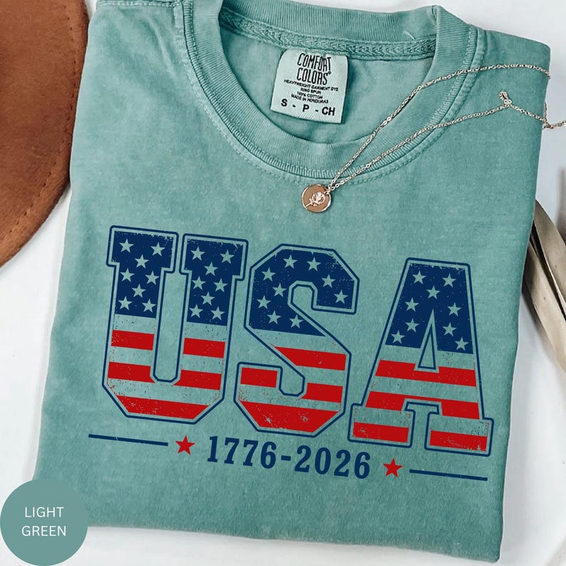A light green t-shirt with the text "USA" and "1776-2026" printed on it, along with an American flag design.
