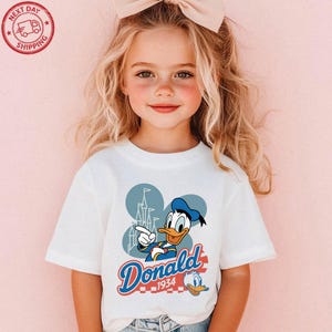 A young girl with blonde hair wearing a white t-shirt with a Disney character printed on it.