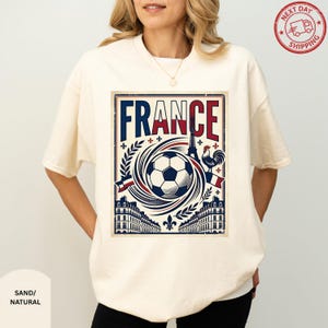 A woman wearing a white t-shirt with a graphic design featuring the word "FRANCE" and various elements associated with the country, such as a soccer ball, buildings, and a rooster.