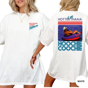 Puede incluir: Camiseta blanca con el texto "WAYDOWN YONDER ON THE Chattahoochee" en la parte delantera. La parte trasera de la camiseta presenta un gr&aacute;fico con el texto "HOTTER THANA" y "HOOCHIE COOCHIE" con una imagen de una persona en un tubo.
