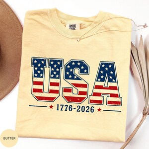 A yellow t-shirt with the text "USA" and "1776-2026" printed on it, along with an American flag design.