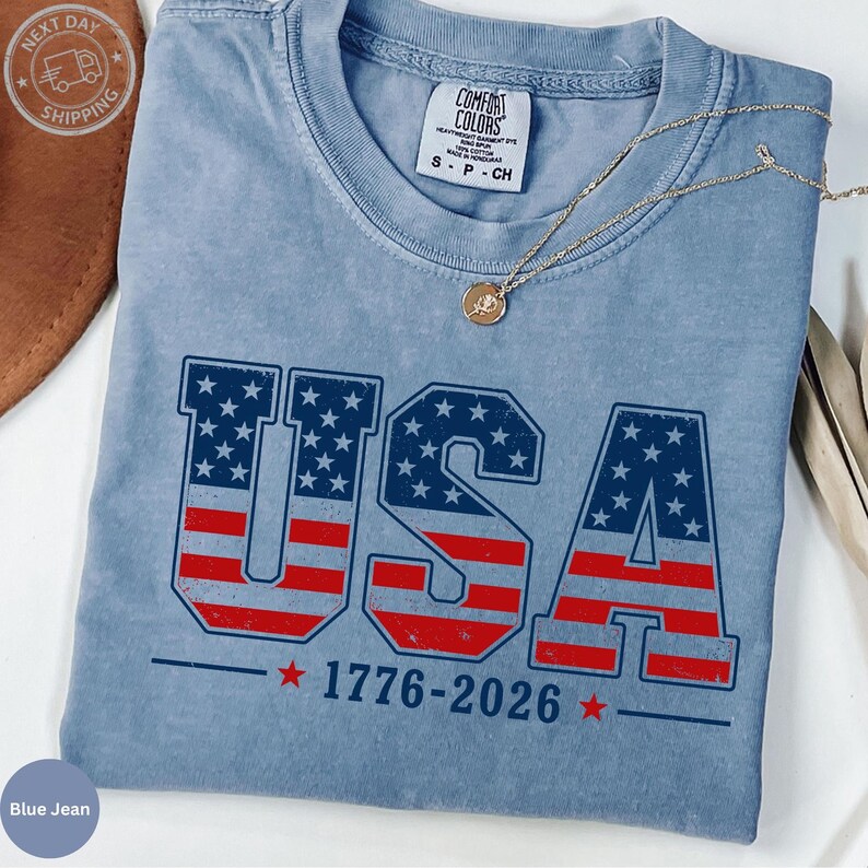 A blue t-shirt with the text "USA" and "1776-2026" printed on it, along with an American flag design. The t-shirt is folded and placed on a white surface, with a necklace and a brown hat visible in the background.