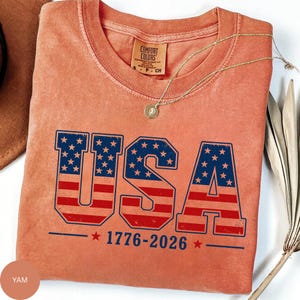A red t-shirt with the text "USA" printed on it, along with the year "1776-2026" below it. The t-shirt is displayed on a white surface, and there is a necklace and some leaves in the background.