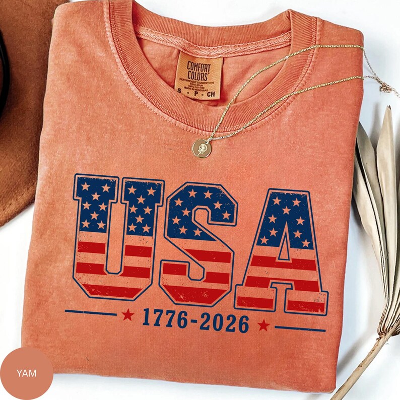 A red t-shirt with the text "USA" printed on it, along with the year "1776-2026" below it. The t-shirt is displayed on a white surface, and there is a necklace and some leaves in the background.