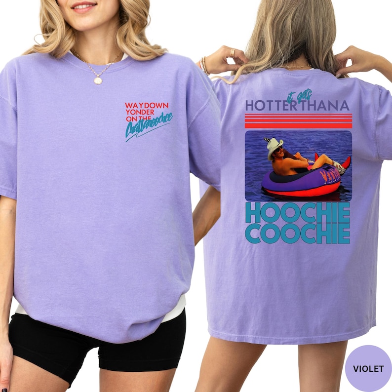 Puede incluir: Camiseta violeta de manga corta con texto en la parte delantera y trasera. La parte delantera dice "WAYDOWN YONDER ON THE Chattahoochee" en una fuente retro. La parte trasera tiene un gr&aacute;fico de una persona en un tubo y las palabras "HOTTER THANA HOOCHIE COOCHIE."