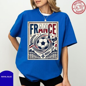 A woman wearing a blue t-shirt with a graphic design featuring a soccer ball, buildings, and a rooster, along with the word "FRANCE" prominently displayed.
