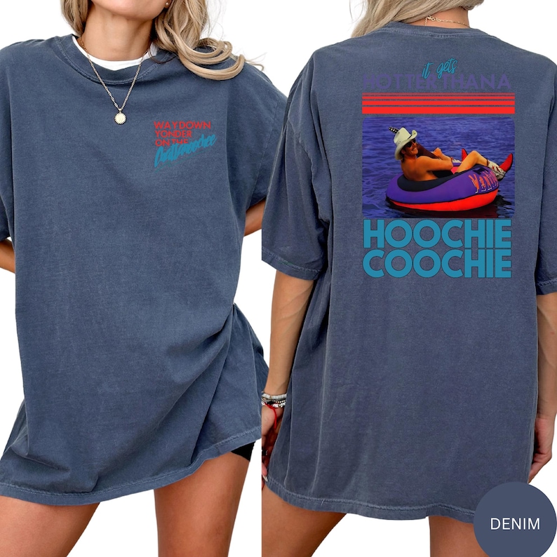 Puede incluir: Camiseta azul denim con el texto "Waydown Yonder Outfitters" en rojo y turquesa en la parte delantera. La parte trasera de la camiseta presenta un gr&aacute;fico de una persona en un flotador con el texto "It gets hotter than a Hoochie Coochie".