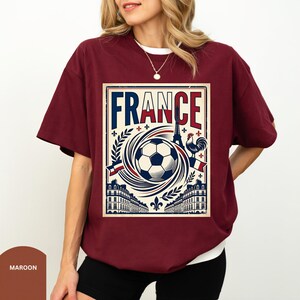 A woman wearing a burgundy t-shirt with a graphic design featuring the word "FRANCE" and various elements associated with the country, such as a soccer ball, buildings, and a rooster.