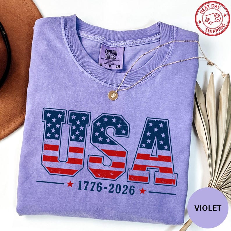 a purple t-shirt with the text "USA" printed on it, along with the numbers "1776-2026" below it. The t-shirt is laid out on a white surface, and there is a brown hat and some dried leaves nearby.