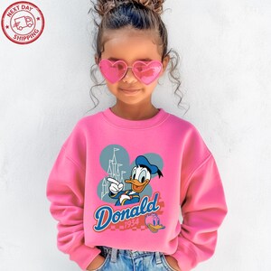 A young girl wearing a pink sweater with a Disney character and heart-shaped sunglasses, smiling at the camera.
