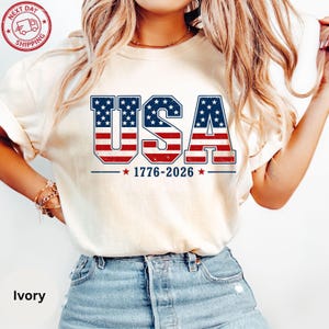 A woman wearing a white t-shirt with the text "USA" printed on it, along with the years 1776-2026.