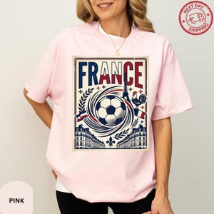 A woman wearing a pink t-shirt with a graphic design of a soccer ball, buildings, and a rooster, along with the word "FRANCE" prominently displayed.