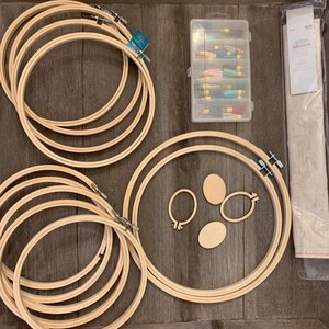 May include: A collection of wooden embroidery hoops in various sizes, along with small oval wooden pieces and a clear plastic container filled with colorful tassels. A piece of fabric is also included, all arranged on a wooden surface.
