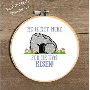May include: A white embroidery hoop with a cross stitch design depicting a stone tomb with the text "He is not here; for he has risen!" in purple lettering. The tomb is surrounded by green grass and pink flowers.