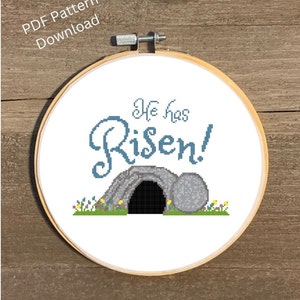 May include: A white embroidery hoop with a cross stitch design of a stone tomb with the words "He has Risen!" in blue thread.