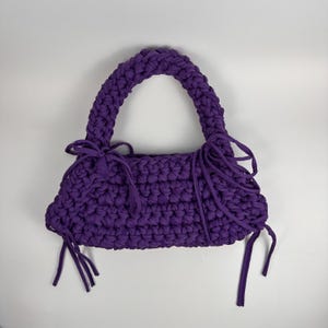 May include: A handmade, purple, crocheted handbag with a rounded handle and decorative fringe. The bag is made of thick, textured yarn and has a rectangular shape. The handle is also crocheted and appears sturdy. The fringe is tied to the sides of the bag.