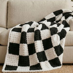 Könnte beinhalten: Eine schwarz-weiß karierte Häkeldecke, die über einem beigefarbenen Sofa drapiert ist. Das Muster der Decke ist ein kühnes, klassisches Schachbrettmuster. Eine Vase mit getrockneten Pflanzen steht auf einem hölzernen Beistelltisch neben dem Sofa. Ein Knäuel weißer Wolle liegt auf dem Boden.