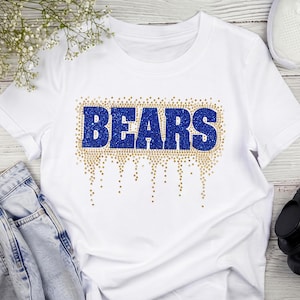 Puede incluir: Una camiseta blanca de manga corta con la palabra BEARS en letras de bloque de color azul brillante. El texto está rodeado por un efecto de goteo decorativo hecho de pequeños puntos dorados brillantes que se extienden hacia abajo.