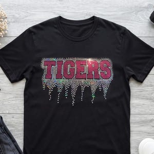 Op de afbeelding: Een zwart t-shirt met korte mouwen en het woord TIGERS in roze strass-steentjes op de borst. De letters hebben een druipend effect, gecreëerd met verspreide doorzichtige en iriserende strass-steentjes die vanaf de tekst naar beneden lopen.