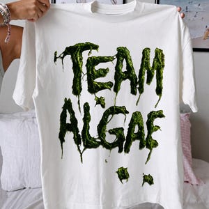 May include: A white short-sleeved t-shirt featuring a graphic print on the front. The text reads "TEAM ALGAE" in a stylized, dripping, textured green font that resembles slimy moss or seaweed, with small green splatters below the text.