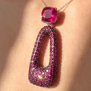 May include: A pendant necklace featuring a large, cushion-cut magenta gemstone set above a larger, elongated, open-frame pendant encrusted with numerous small, round, deep pink stones set in a dark, metallic frame on a thin silver-toned chain.