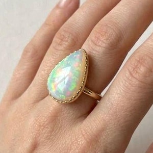 Könnte beinhalten: Ein goldener Ring mit einem großen, tropfenförmigen Opal-Edelstein. Der Opal zeigt schillernde Farben und ist in einer Goldfassung mit einem Perlenrand gefasst. Der Ring wird an einer Hand gezeigt.