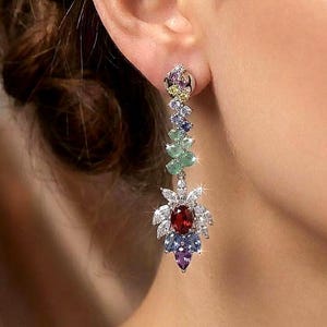 Könnte beinhalten: Ein langer, baumelnder Ohrring mit floralem Design. Der Ohrring hat eine Ansammlung farbiger Edelsteine, darunter Rot, Blau, Grün und Lila, die in einem silbernen oder weißen Metall gefasst sind. Der Ohrring wird am Ohr getragen.