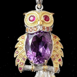 Könnte beinhalten: Ein Eulenanhänger aus Gold und Silber an einer Goldkette. Die Eule hat einen großen ovalen violetten Amethyst auf der Brust, zwei runde rote Rubinaugen und Flügel, die mit kleinen orangefarbenen und roten Edelsteinen besetzt sind.