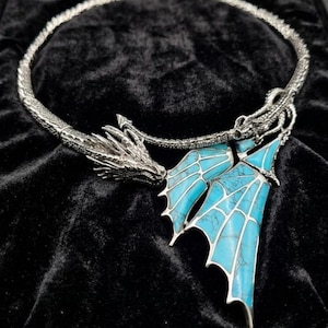 May include: A silver-toned metal necklace featuring a detailed dragon head at one end and a pair of articulated, bright turquoise wing-shaped pendants with dark veining patterns attached to the other side of the circular neck band.