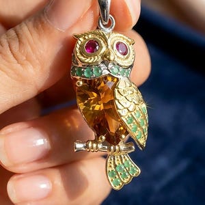 May include: An owl-shaped pendant featuring a large, faceted amber-coloured gemstone for the body, two round red gemstone eyes, and small green gemstones embedded along the neck, wing, and tail. The owl is perched on a silver-toned metal branch.