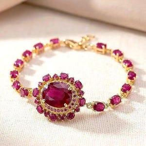 Puede incluir: Pulsera dorada con un gran rubí ovalado rodeado de rubíes más pequeños. La cadena de la pulsera está adornada con rubíes ovalados adicionales, creando un diseño lujoso y elegante. Una hermosa joya.