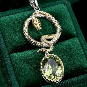 Puede incluir: Un colgante que presenta una serpiente texturizada y enroscada en tonos dorados y plateados, formando una figura circular. Debajo de la serpiente, una piedra preciosa ovalada de color verde claro facetada está engastada en un marco de metal festoneado.