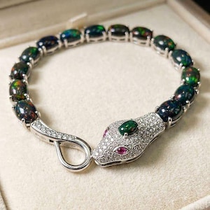 May include: A snake-shaped bracelet with oval-cut black opals and a silver clasp. The snake's head is encrusted with small diamonds, a green emerald, and two small pink gemstones. The bracelet is displayed in a beige jewelry box.