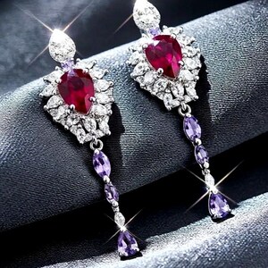 May include: A pair of drop earrings featuring a central pear-shaped red gemstone surrounded by a cluster of clear white stones. A dangling chain extends from the base, adorned with three small, oval-shaped purple gemstones in a vertical arrangement.