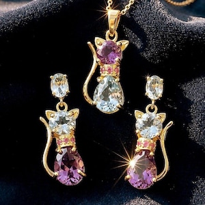 以下が含まれることがあります： 金色の猫のデザインのジュエリーセット。セットには、ペンダントネックレスとマッチするイヤリングが含まれています。各作品は、紫、ピンク、青の宝石が付いた猫のシルエットです。ジュエリーは、暗くテクスチャ加工された表面に表示されています。