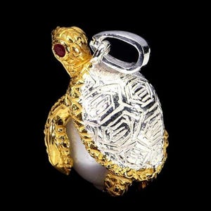 Puede incluir: Un colgante de tortuga de oro y plata con un ojo de piedra preciosa roja. El colgante tiene un diseño detallado y texturizado y una fianza plateada para sujetarlo a una cadena. El caparazón de la tortuga es plateado, y la cabeza y las patas son doradas.