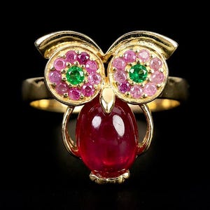 May include: A gold ring shaped like an owl, featuring a large cabochon ruby body, two circular eyes set with green centre stones surrounded by pink sapphires, and a small gold beak. The band is smooth gold, and the design is symmetrical.