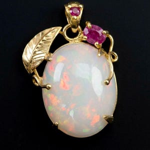 May include: A gold pendant featuring a large oval cabochon opal with iridescent flashes of red, green, and orange. The setting includes a textured gold leaf accent on the left and two small, round, deep pink rubies set near the top bail.