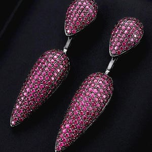 Könnte beinhalten: Ein Paar Ohrhänger, bestehend aus jeweils zwei tropfenförmigen Elementen, die vollständig mit kleinen, leuchtend pinkfarbenen Steinen in einer dunklen Metallfassung besetzt sind. Das Design umfasst einen kleineren oberen Tropfen und einen größeren, länglichen unteren Tropfen.