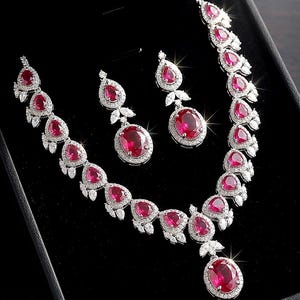 Puede incluir: Un conjunto de joyería que incluye un collar y pendientes a juego con piedras rojas vibrantes de forma ovalada y de pera. Cada pieza está montada en un marco de metal plateado incrustado con pequeñas piedras claras y brillantes.