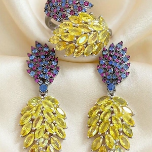 May include: A jewelry set consisting of a ring and two matching drop earrings. Each piece features a cluster of marquise-cut yellow stones at the base, topped with a smaller section of round-cut pink and blue stones set in dark metal.