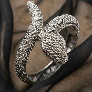 May include: A silver-toned snake bracelet with intricate detailing. The snake's head features a small red gemstone for the eye. The bracelet is displayed on a black and brown fabric background, showcasing the jewelry's design.