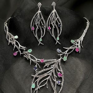 Puede incluir: Un conjunto de joyería en tono plateado que consta de un collar y pendientes colgantes a juego. Las piezas presentan un diseño de ramas incrustado con pequeñas piedras claras y acentuado con gemas ovaladas rosas, verdes y azul oscuro.