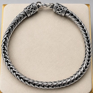 May include: A silver-colored braided chain necklace. The necklace features a thick, woven design with decorative end pieces and a clasp. The ends of the necklace have a detailed pattern. The necklace is displayed on a neutral-toned surface.