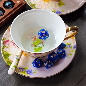 May include: A white teacup with a gold handle and floral design, filled with water and a blue twenty-sided die. The cup sits on a matching saucer with additional blue dice. A wooden box with dice is in the background.