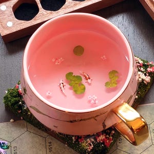 May include: A pink ceramic mug with a gold handle, filled with pink liquid and miniature koi fish, lily pads, and flowers. The mug sits on a floral coaster, with a wooden tray in the background.