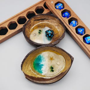May include: Two coconut shell bowls with resin art depicting a beach scene, with a wooden dice tray. The bowls have a white and turquoise resin design with small embellishments. A black and blue twenty-sided die rests in one bowl.