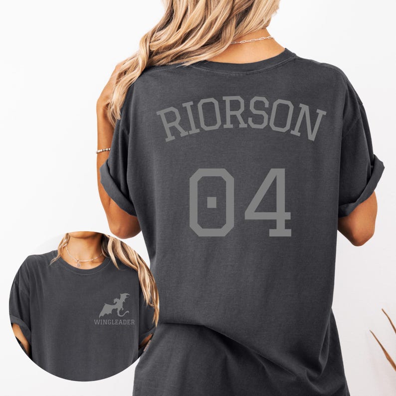 May include: A charcoal gray t-shirt featuring the text RIORSON and the number 04 in a large athletic font on the back. The front chest area displays a small graphic of a dragon silhouette above the text WINGLEADER in a simple sans-serif font.