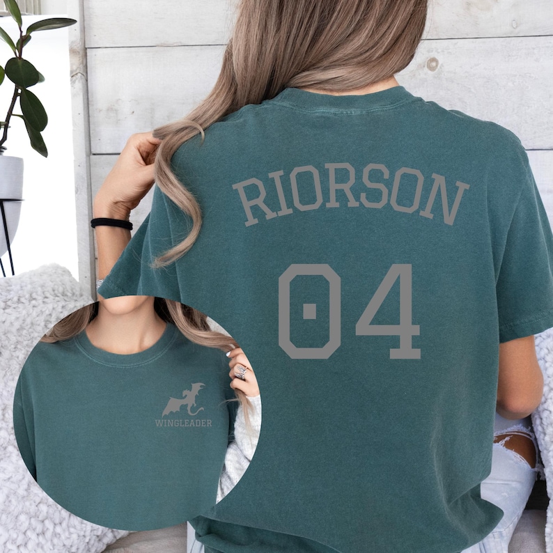 May include: A dark teal short-sleeved t-shirt featuring the text RIORSON and the number 04 in a grey athletic font on the back. The front chest area displays a small grey graphic of a dragon silhouette above the text WINGLEADER.